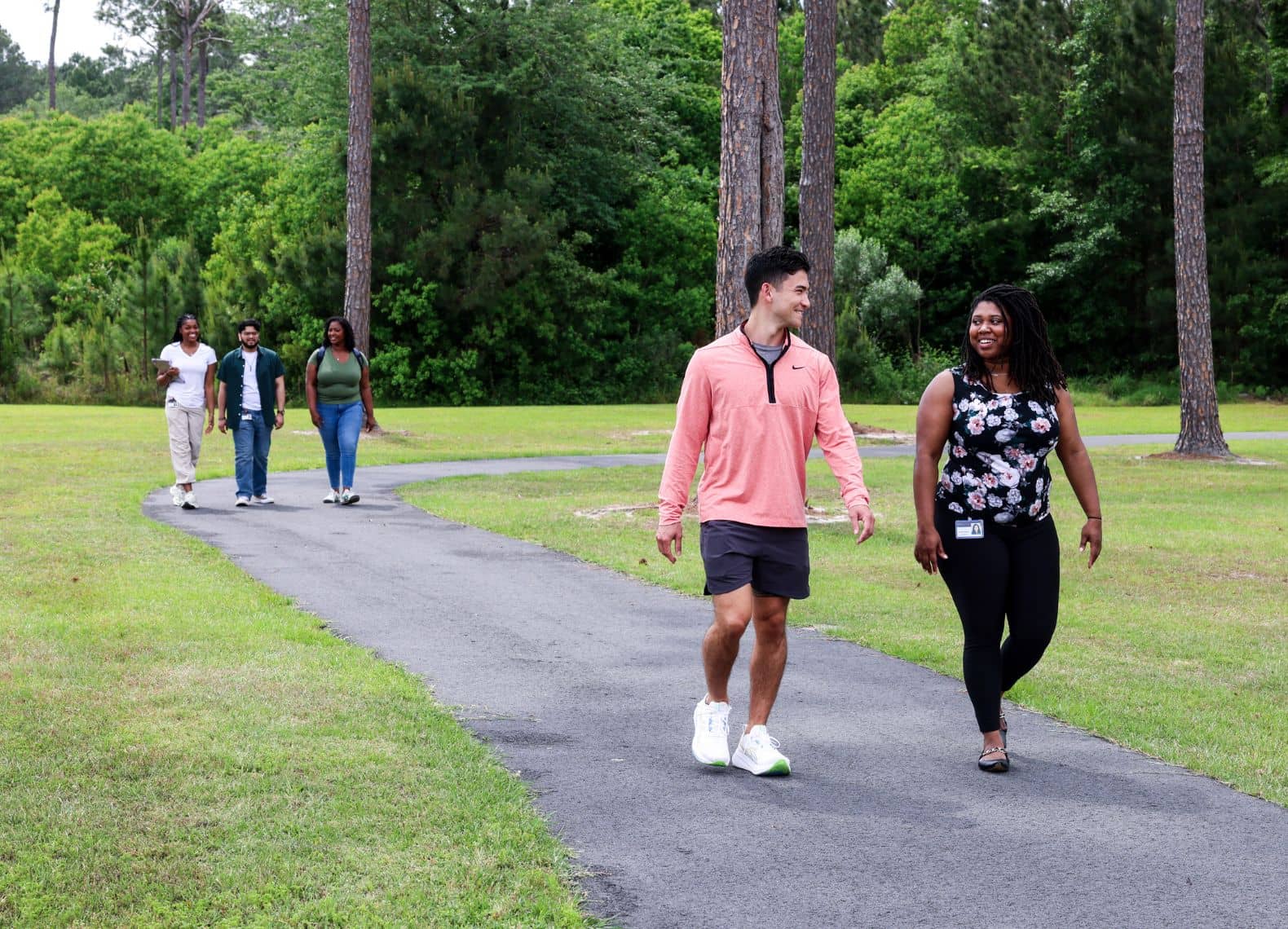Five students walk along a paved path through a green, tree-lined campus lawn