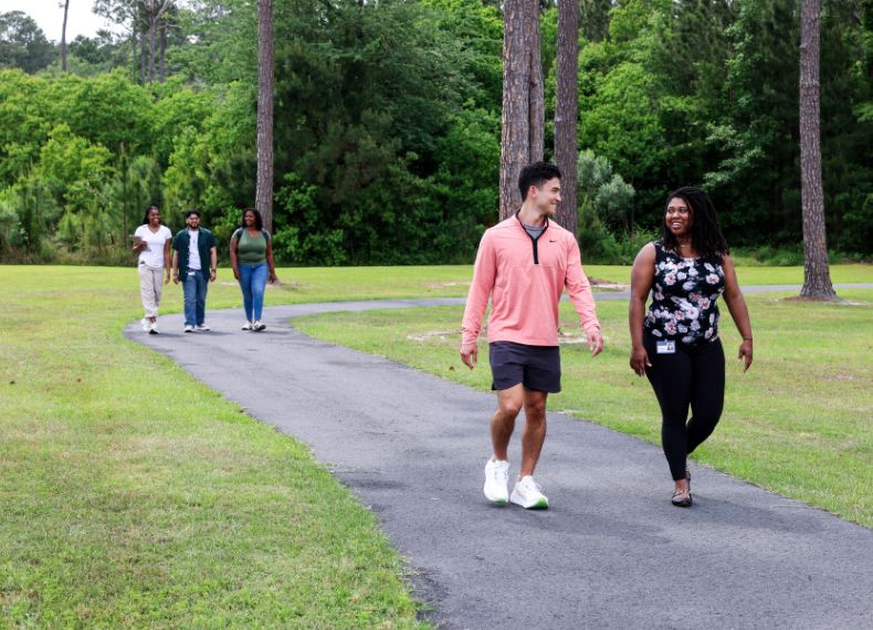 Five students walk along a paved path through a green, tree-lined campus lawn