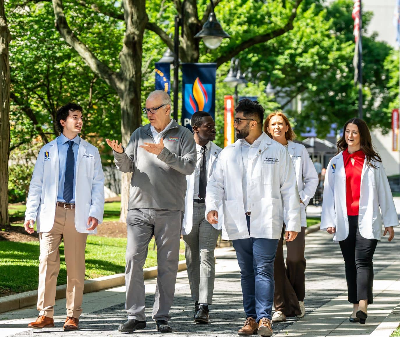 A group of student physicians in white coats walk and talk with a faculty member along a tree-lined PCOM campus walkway with banners