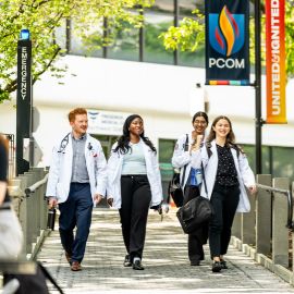 Four students in white coats walk together across a campus bridge with the PCOM building in the background