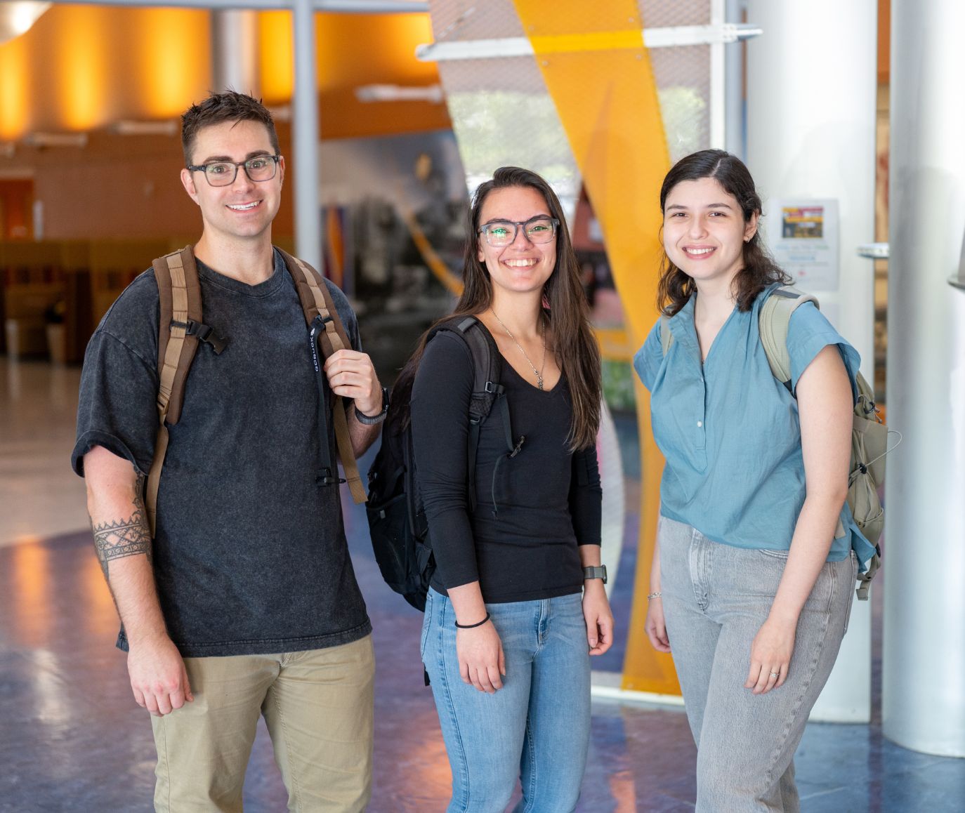 Three students wearing backpacks smile together in a modern campus lobby with colorful architectural details