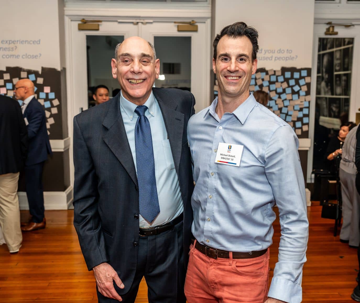 Two men smiling together at a PCOM alumni networking event with sticky note boards in the background