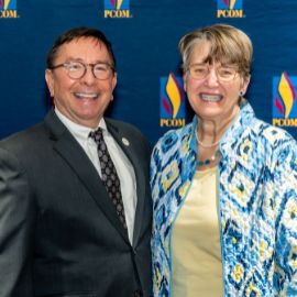 A man in a dark suit and a woman in a blue patterned jacket smile in front of a PCOM branded backdrop