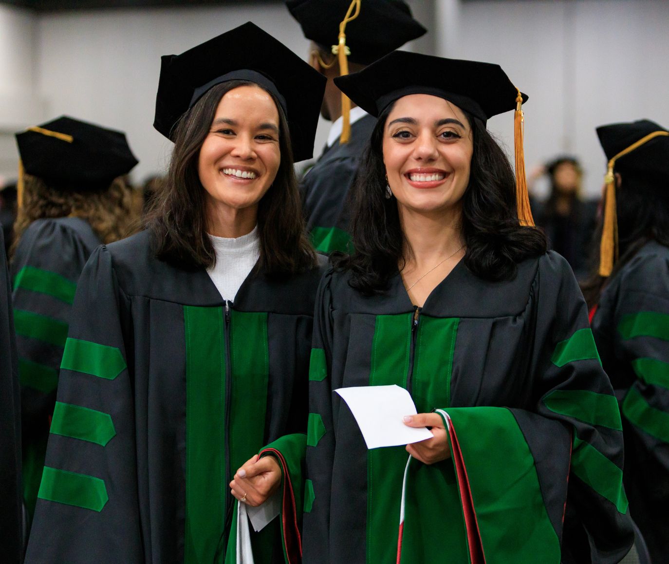 Two smiling graduates in black and green doctoral caps and gowns at a PCOM commencement ceremony
