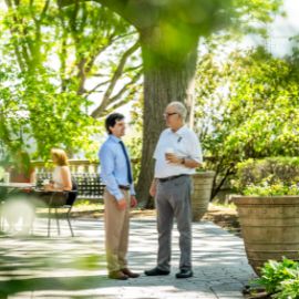Two men stand and talk on a shaded campus garden walkway surrounded by trees and greenery