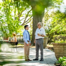 Two men stand and chat on a sunlit stone walkway surrounded by lush campus greenery