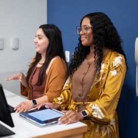 Two students smile while working together with a tablet at a computer station in a campus lab