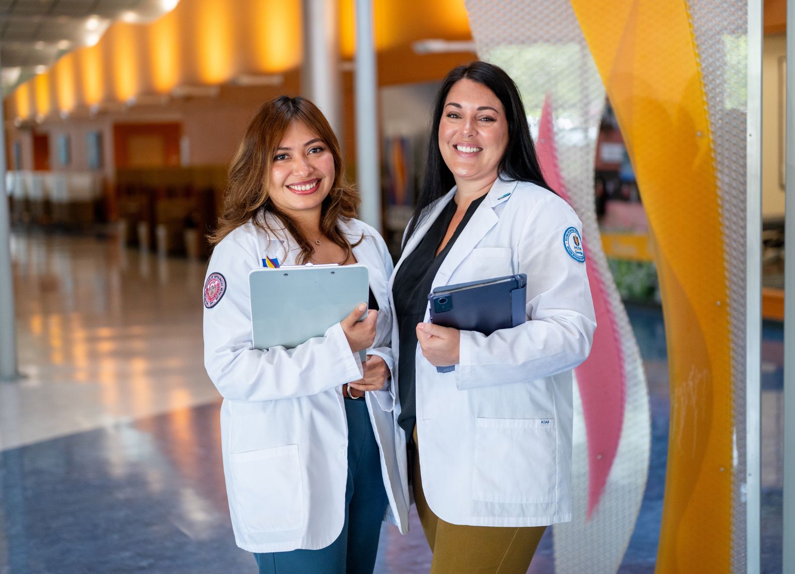 Two smiling students in PCOM white coats hold a clipboard and tablet in a colorful campus hallway