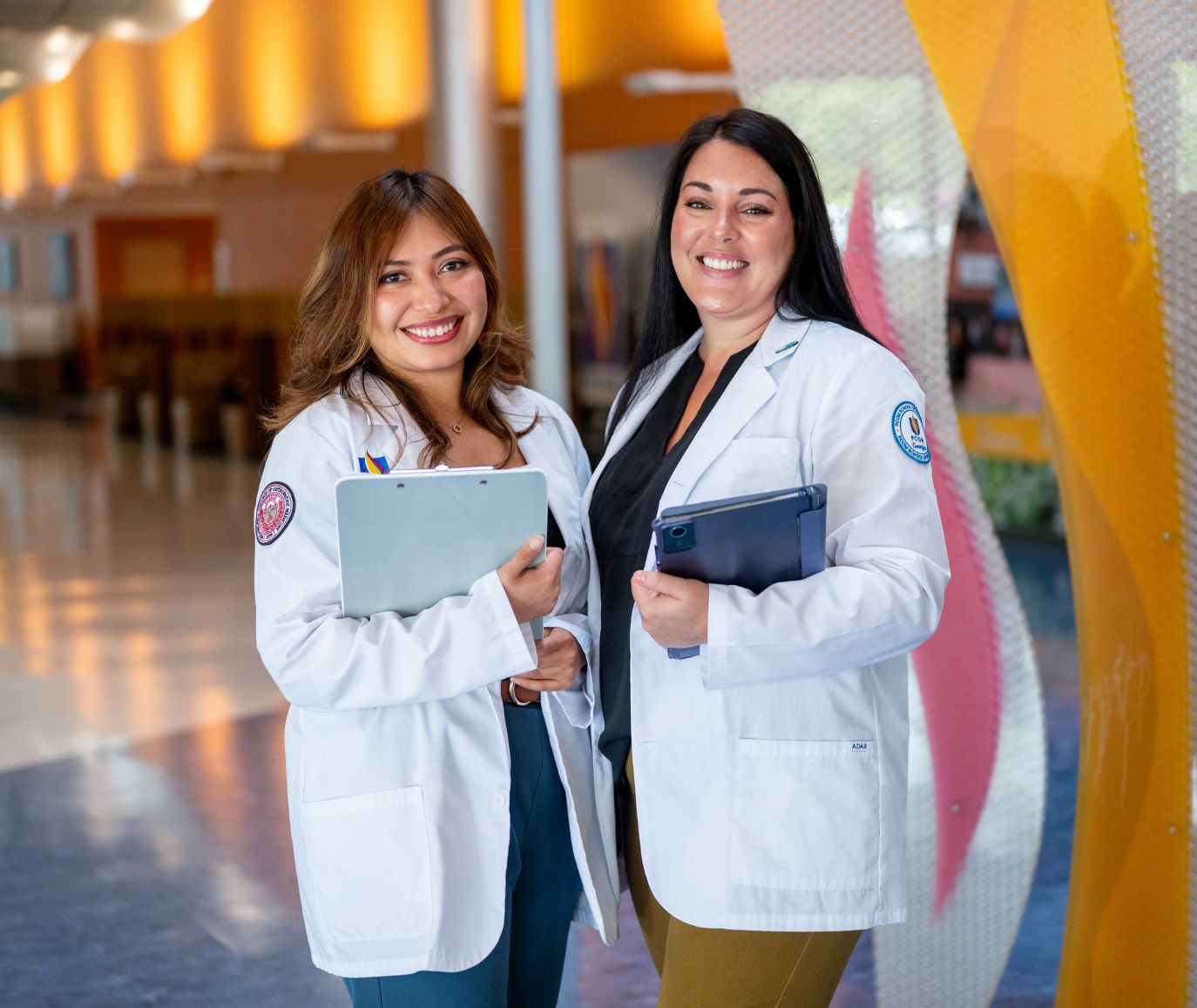 Two smiling students in PCOM white coats hold a clipboard and tablet in a colorful campus hallway