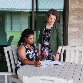 Two women review documents together at an outdoor patio table on a modern campus