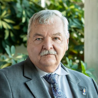 Man with gray hair and a mustache wearing a gray suit and blue paisley tie with a PCOM lapel pin