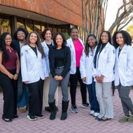 A group of ten women, several wearing white coats, pose together outside a brick campus building